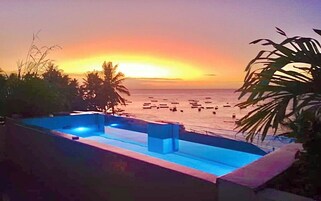 A pool with a sunset view, palm trees, and boats on the water.