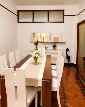 Un coin repas avec une longue table, des chaises blanches, un vase de fleurs et une horloge au mur.