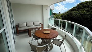A balcony with a round table and chairs, a sofa, and a glass railing overlooking a lush green landscape.