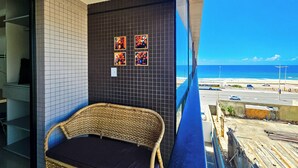 Een balkon met een rieten stoel, een betegelde muur met kunst, en uitzicht op strand en zee.