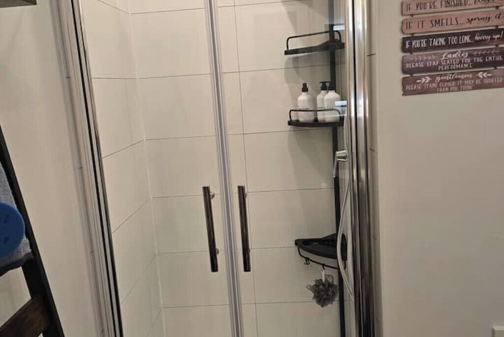 Eine Duschkabine mit Glastür, ein Ablagefach mit Toilettenartikeln und ein Schild mit witzigem Text.