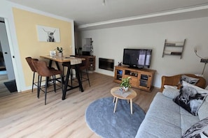 A modern living room with a sofa, a coffee table, a TV, a dining area with chairs, and a small plant.