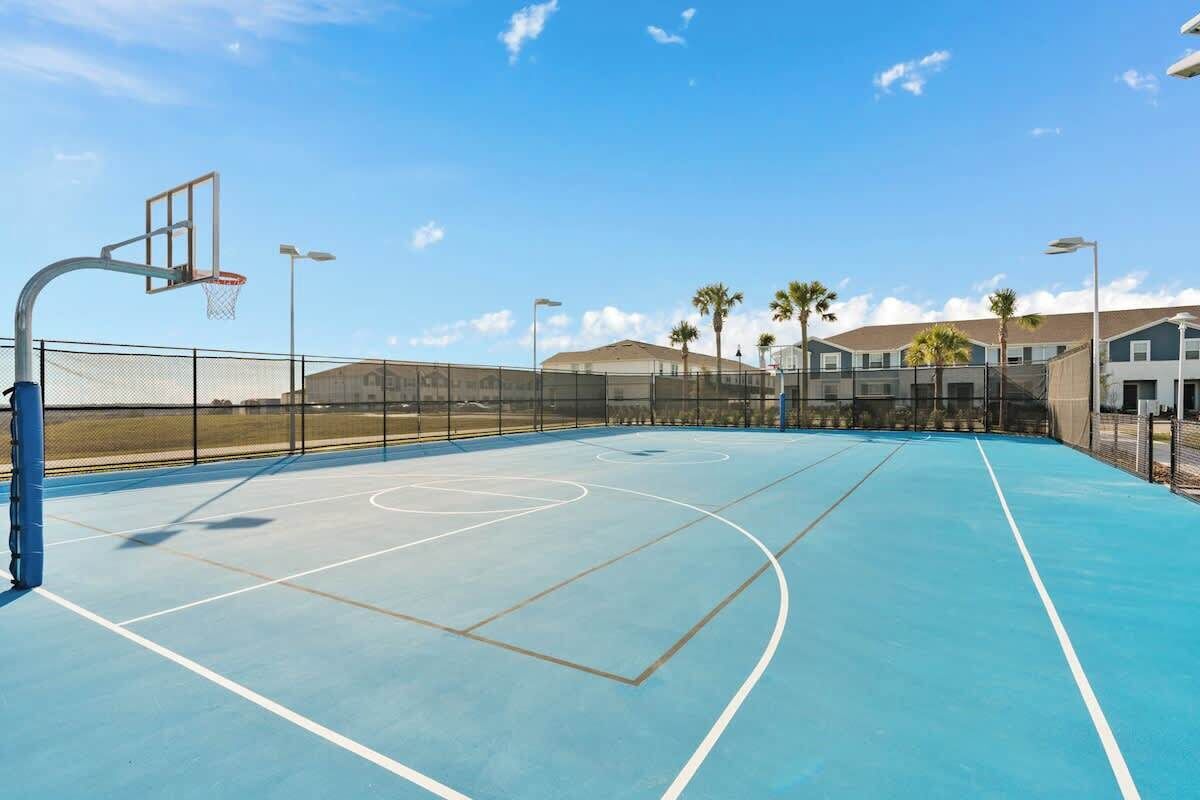 Een buitenbasketbalveld met een blauwe ondergrond, omgeven door een hek, en gebouwen op de achtergrond.