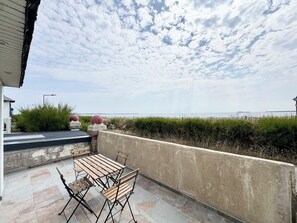 A patio with a table and chairs, a view of the sea, and a hedge in the background.