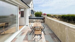 Un patio con una mesa y sillas de madera, un vidrio que da a una habitación y vista al mar.