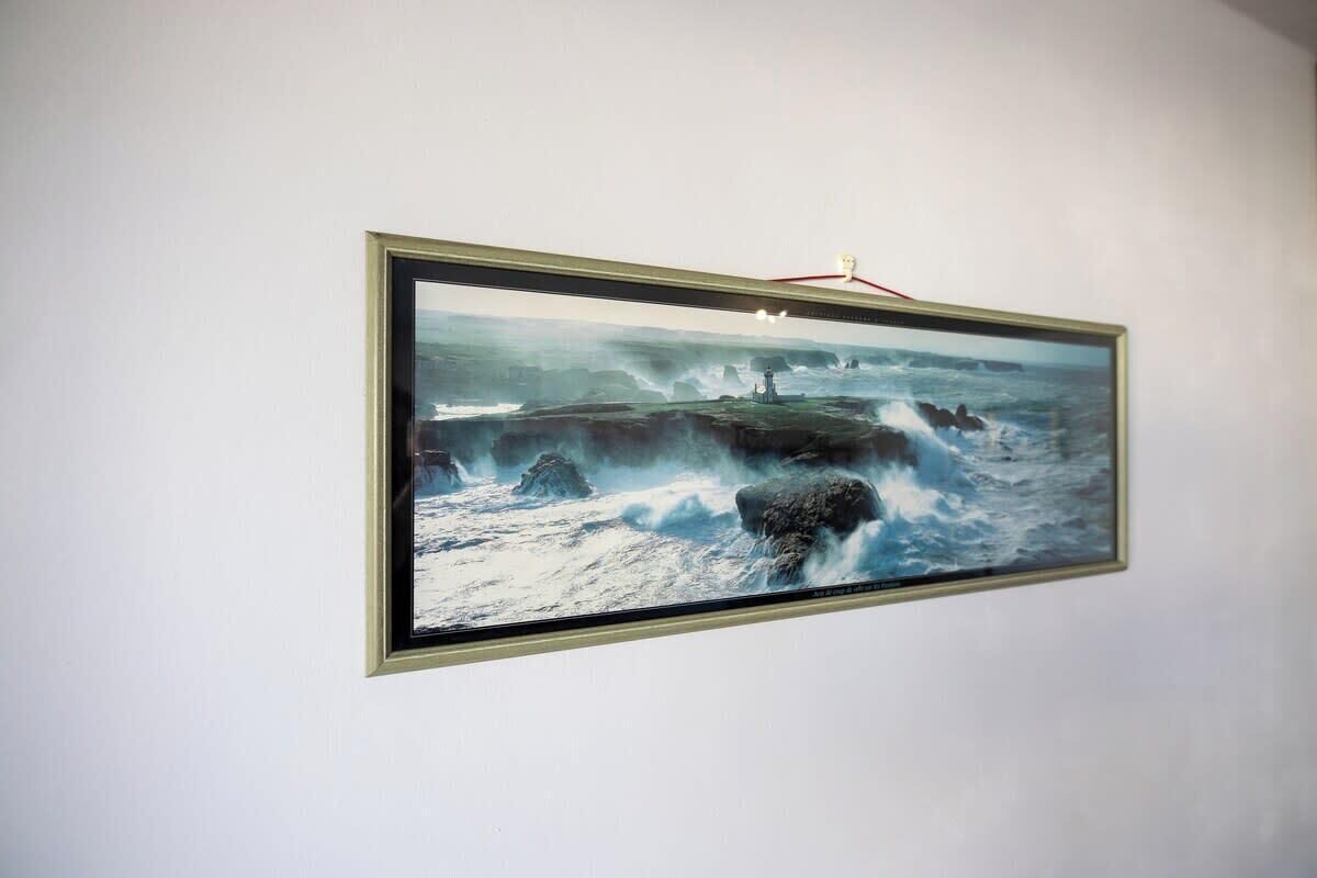 Een lijstwerk met een afbeelding van een zeegezicht, waarop golven tegen rotsen slaan, bevestigd aan een effen muur.