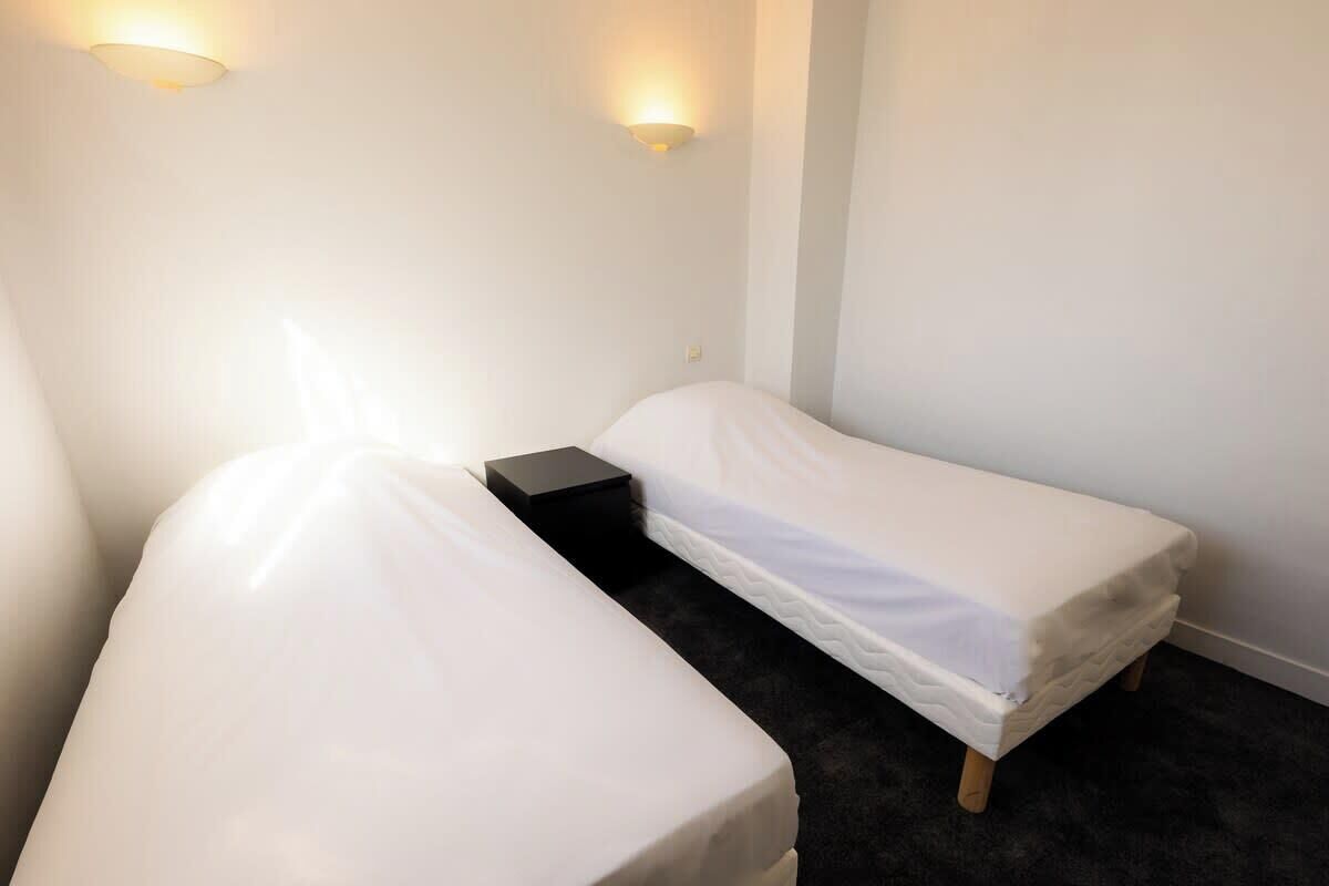 A small, single-room hotel room with two single beds, a bedside table, and two wall-mounted light fixtures.