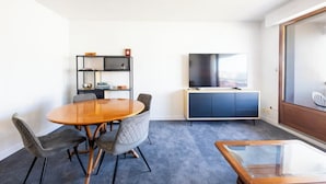 A modern room with a round wooden table, grey upholstered chairs, a bookshelf, a flat-screen TV, a dark blue cabinet, and a glass coffee table. - Plages & bourg à pieds : appartement 2 chambres (Larmor-Plage)