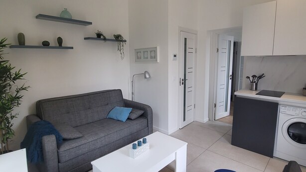 Een moderne woonkamer met een grijze bank, een witte salontafel en een blauwe poef. Er is een keuken met een wasmachine en een aanrecht.