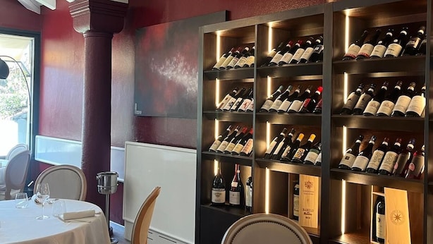 Un interior de restaurante con mesas redondas, sillas, una estantería para vinos y una planta en maceta.
