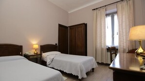 Habitación de hotel con dos camas individuales, un armario de madera, un escritorio con lámpara y una ventana con cortinas.