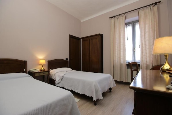 A hotel room with two single beds, a wooden wardrobe, a desk with a lamp, and a window with curtains. - Ristorante Hotel Il Giardinetto (Vercelli)
