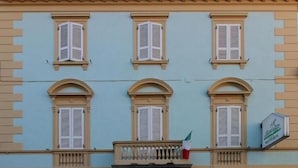 Un edificio con fachada azul claro, cuatro ventanas con contraventanas, un balcón con barandilla y un letrero que indica un restaurante.