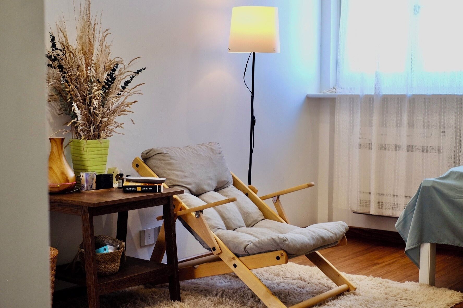 Una habitación con una mecedora de madera, una mesita con libros y un florero, una lámpara de pie y un ventanal con cortinas.