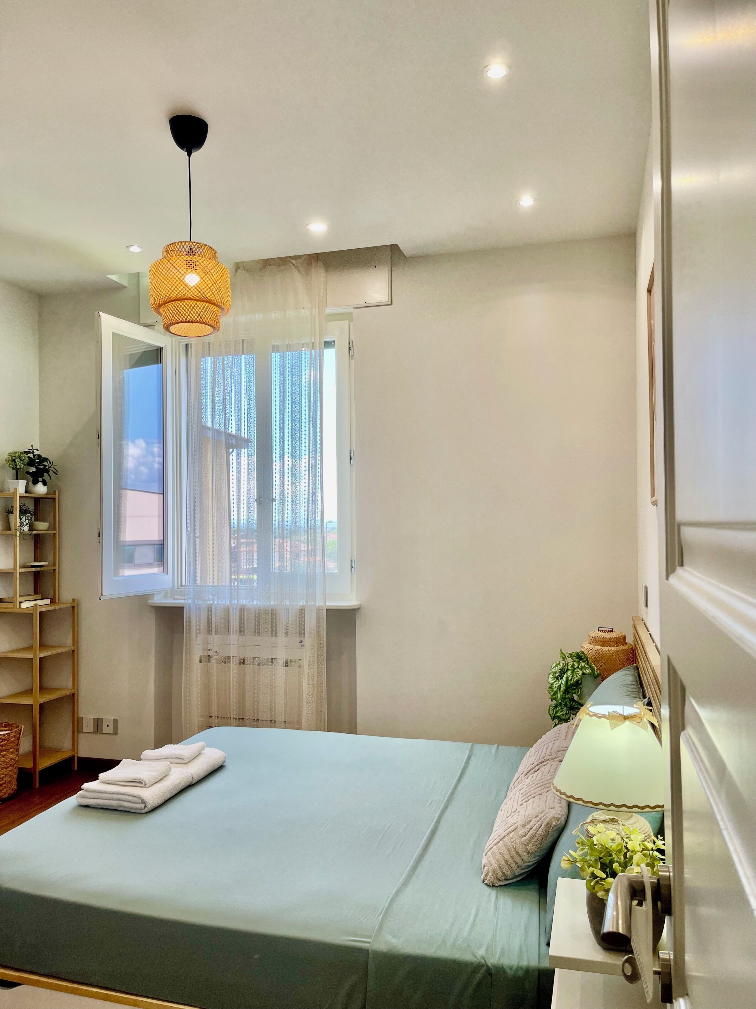 A bedroom with a bed, a bedside table with a lamp, a window with sheer curtains, and a ceiling light fixture.