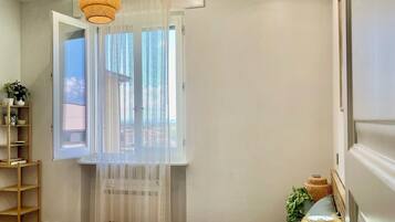 A bedroom with a bed, a bedside table with a lamp, a window with sheer curtains, and a ceiling light fixture.
