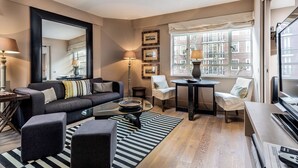 A modern living room with a sofa, ottomans, a glass coffee table, a side table, a lamp, a chair, a small table, and a large window with a view of buildings. - Nell Gwynn House Apartments & Rooms (London)