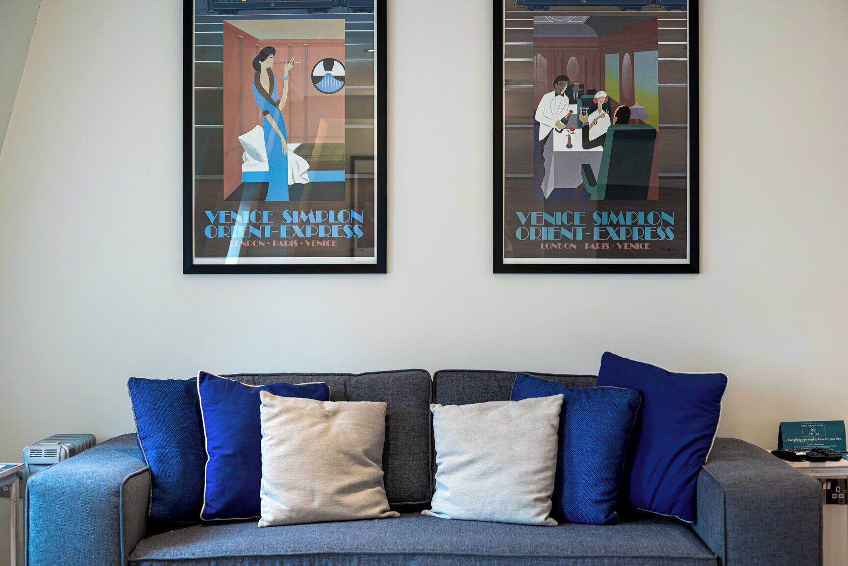 Un canapé gris agrémenté de coussins bleus et beige, deux affiches encadrées représentant le Venice Simplon Orient-Express, et une petite table sur laquelle repose une télécommande.