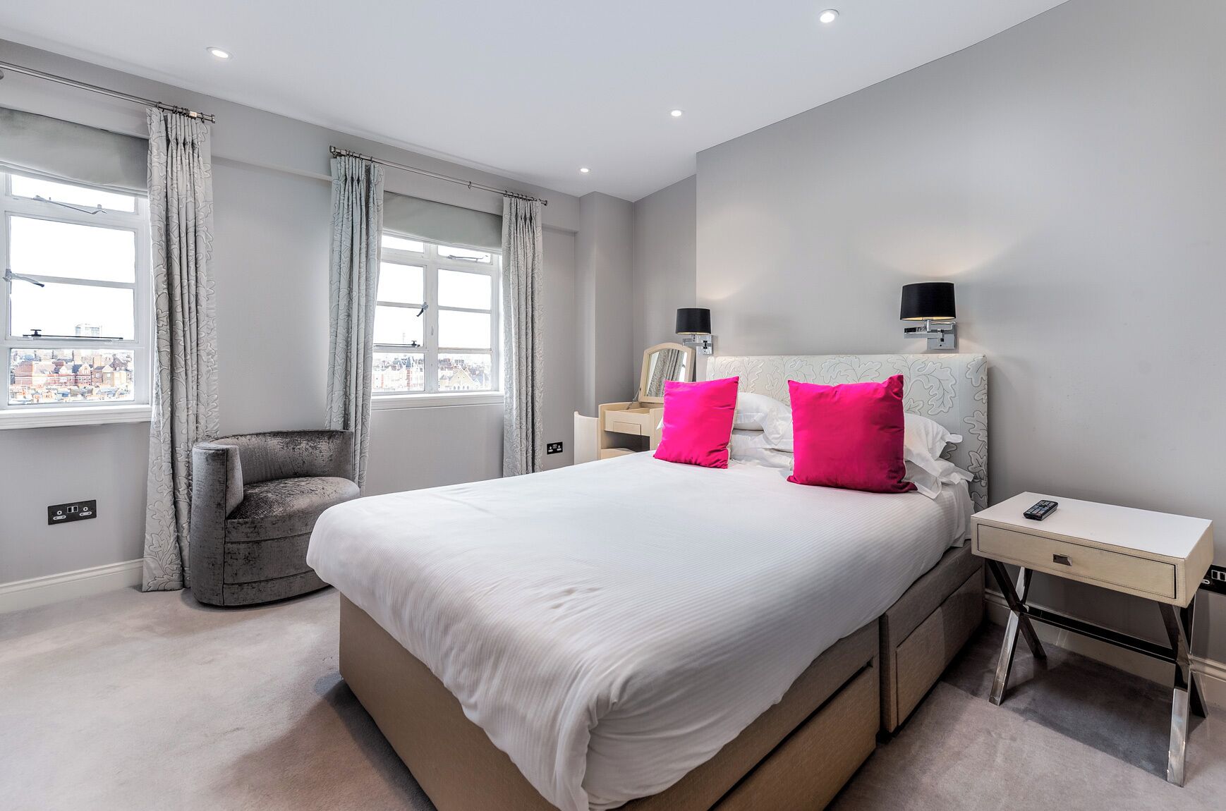 A modern bedroom with a large bed, two bedside tables, a chair, and a view of the city through the window.