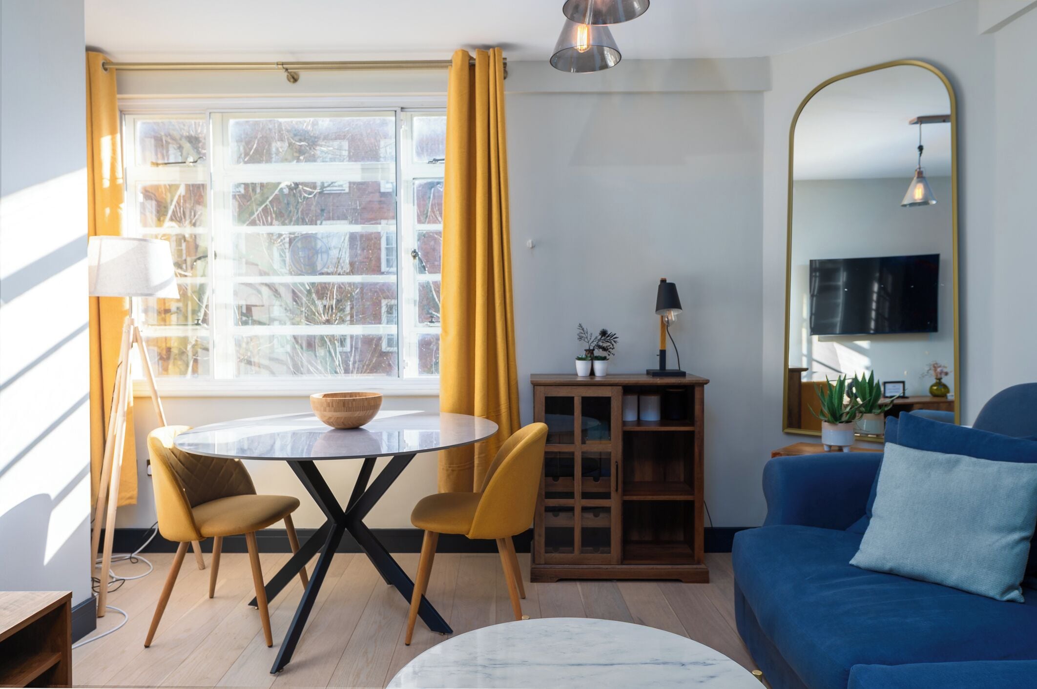 Een woonkamer met een blauwe bank, een ronde tafel met twee stoelen, een houten kast en een groot raam met gele gordijnen.