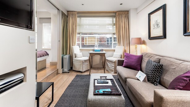 A compact living room with a sofa, a small table, a chair, a TV, and a window with curtains. - Nell Gwynn House Apartments & Rooms (London)
