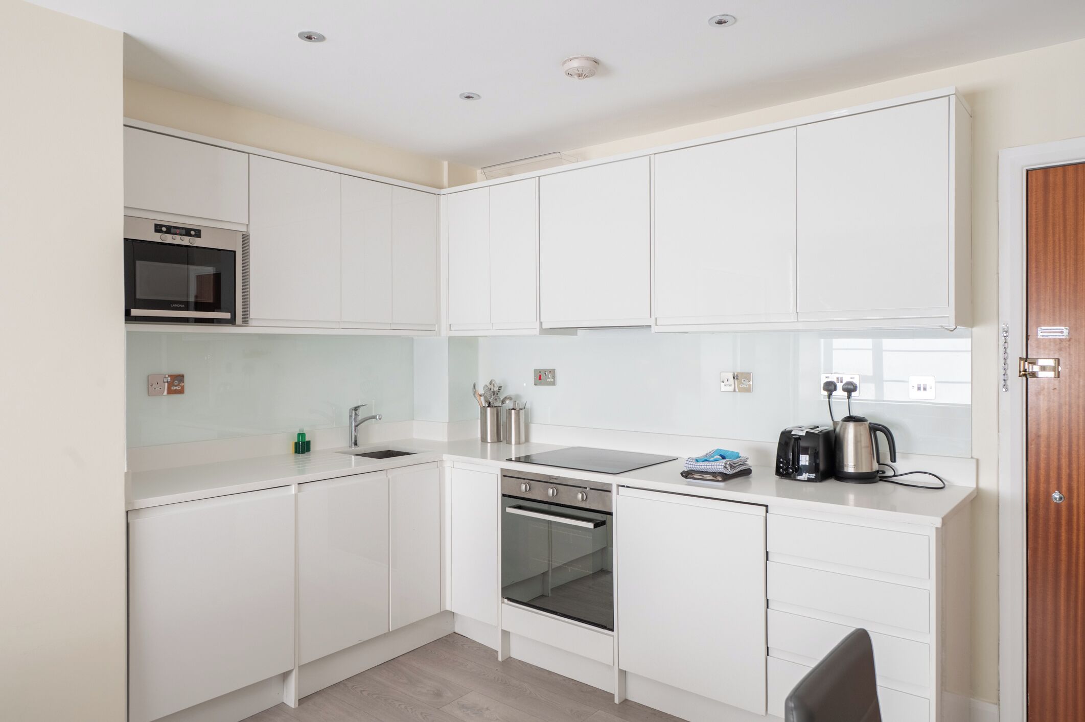 A modern kitchen with white cabinetry, a built-in oven, a microwave, a sink, and a kettle on the countertop.