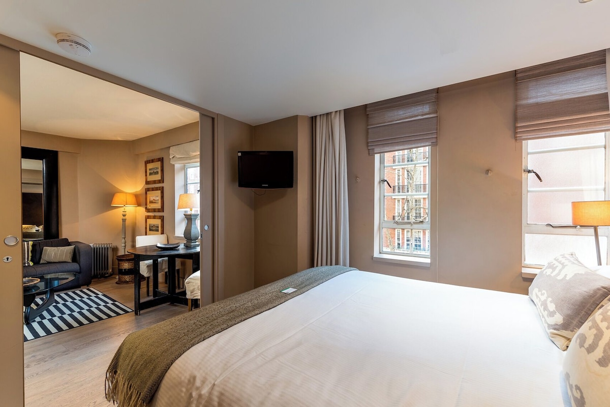 A hotel room with a bed, a television, a desk with a chair, a lamp, and a window with curtains.