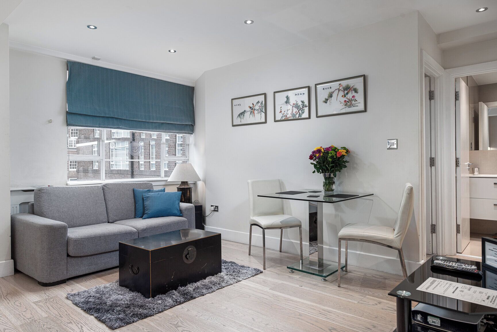 A modern living room with a grey sofa, a glass coffee table, a dining table with chairs, and artwork on the wall.