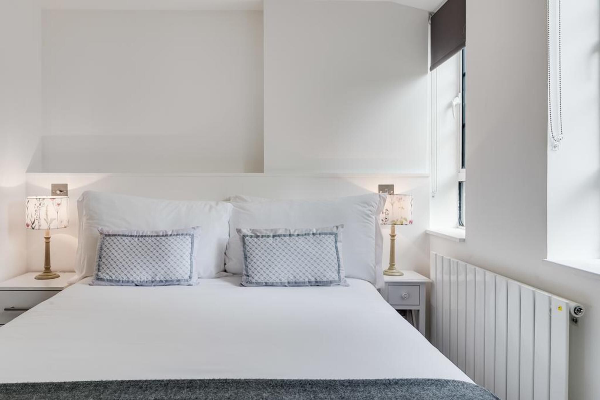 A neatly made bed with white bedding and two patterned pillows, a bedside table with a lamp, and a window with a view of the outside.