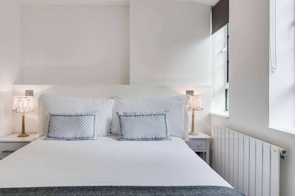 A neatly made bed with white bedding and two patterned pillows, a bedside table with a lamp, and a window with a view of the outside.