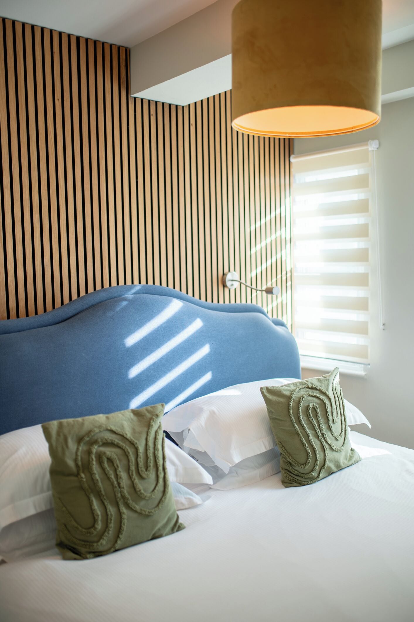 A bed with a blue headboard, two pillows, a wall with vertical wooden slats, and a window with blinds.