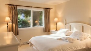 A neatly made bed with white linens, a bedside table with a lamp, and a window with curtains.