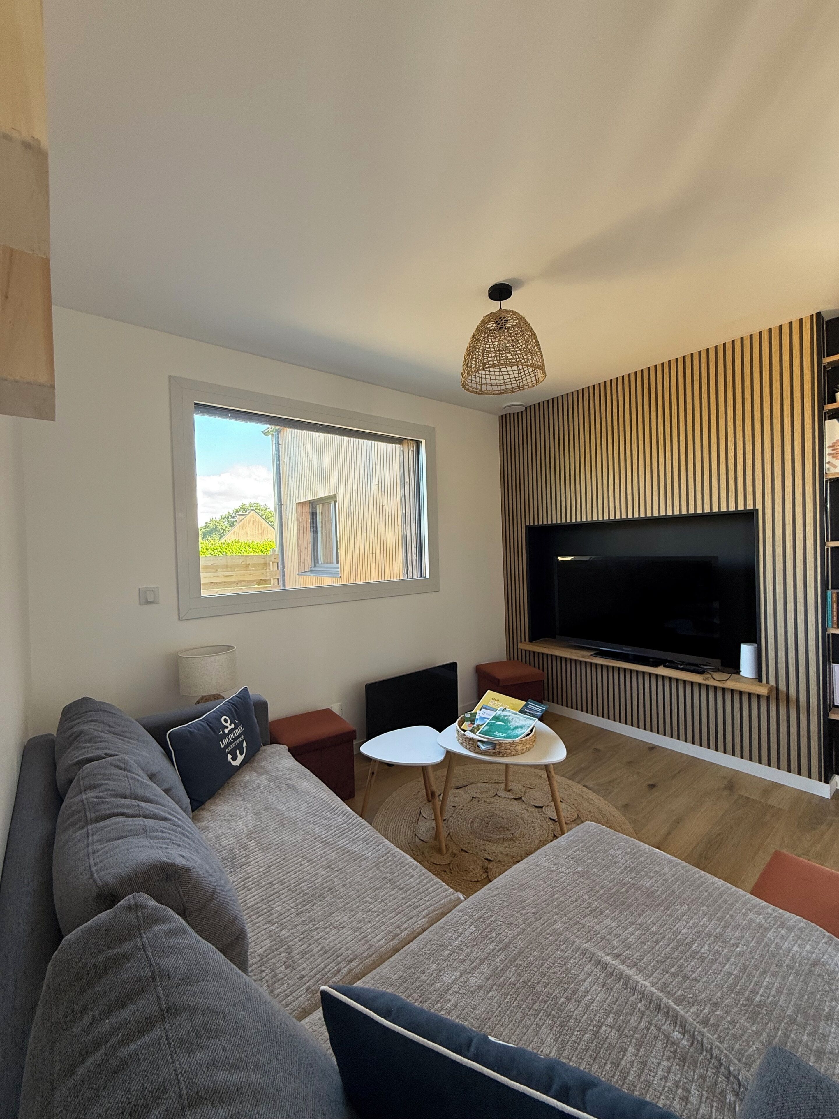 A modern living room with a large flat-screen TV, a sofa, a coffee table, and a window with a view of greenery.
