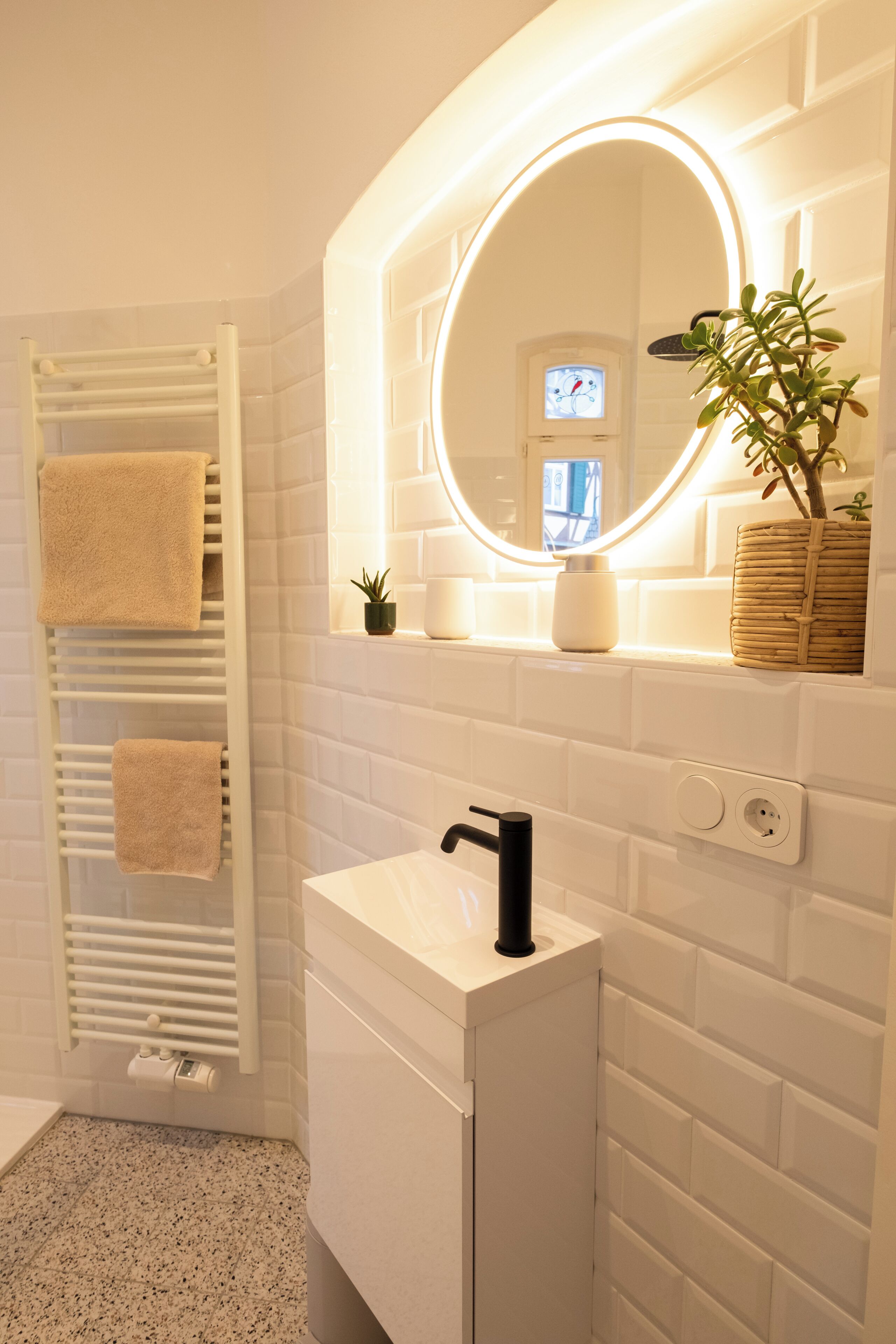 A bathroom with a white tiled wall, a white sink with a black faucet, a towel rack with two towels, a small potted plant, and a mirror with a light.