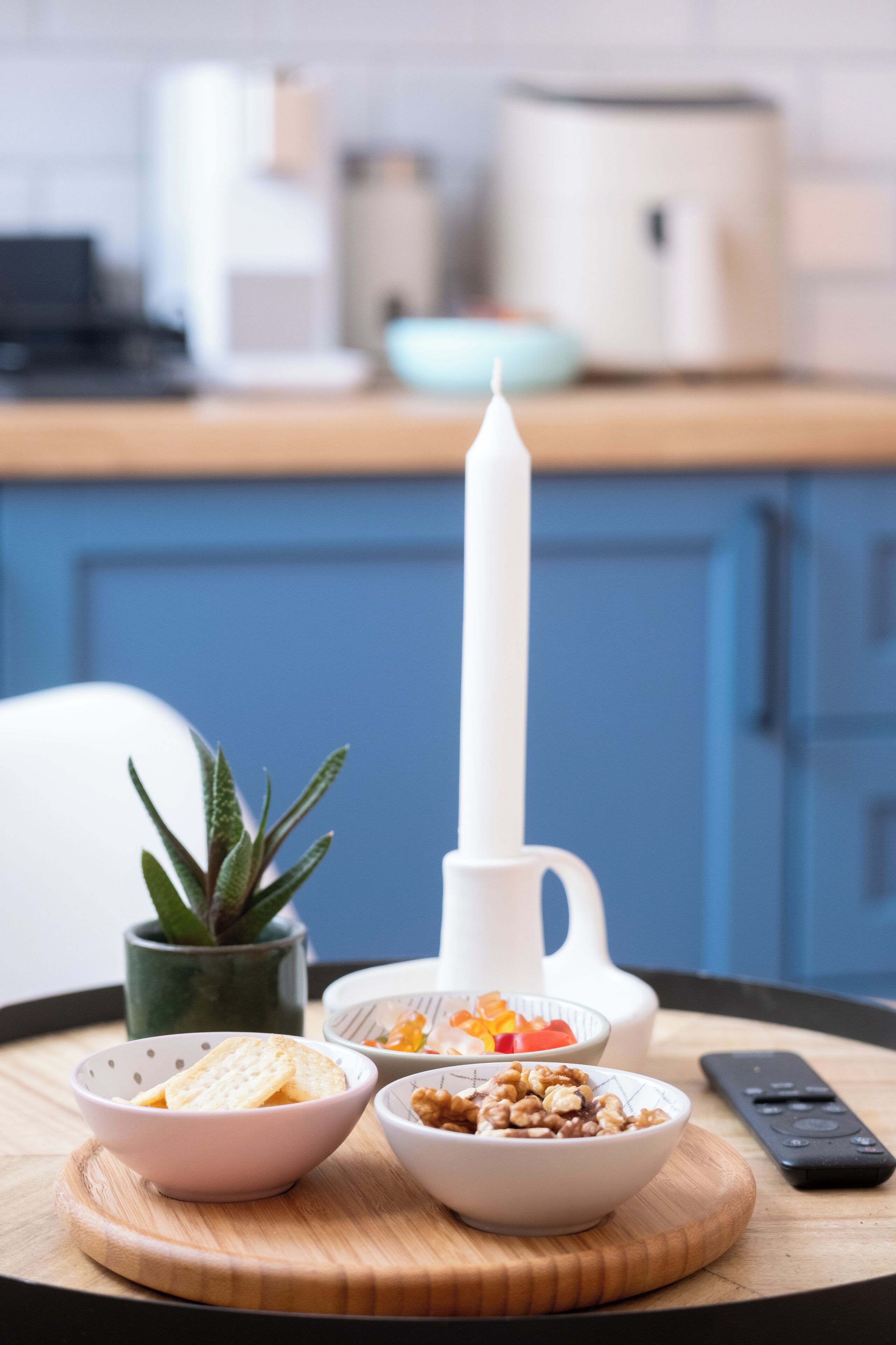 Una encimera de cocina con una vela blanca, un tazón de cereal, un tazón de nueces, un control remoto y una planta en maceta.