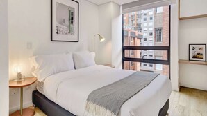 A modern bedroom with a bed, bedside table, lamp, framed picture, and a window with a view of buildings. - w* | Soft Light Loft in the Heart of Bogotá (Bogotá)