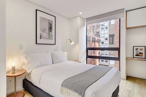 A modern bedroom with a bed, bedside table, lamp, framed picture, and a window with a view of buildings. - w* | Soft Light Loft in the Heart of Bogotá (Bogotá)