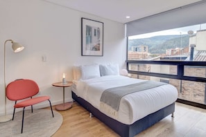 Een moderne slaapkamer met een groot bed, een rode stoel, een nachtkastje met een lamp en een uitzicht op gebouwen door een groot raam.