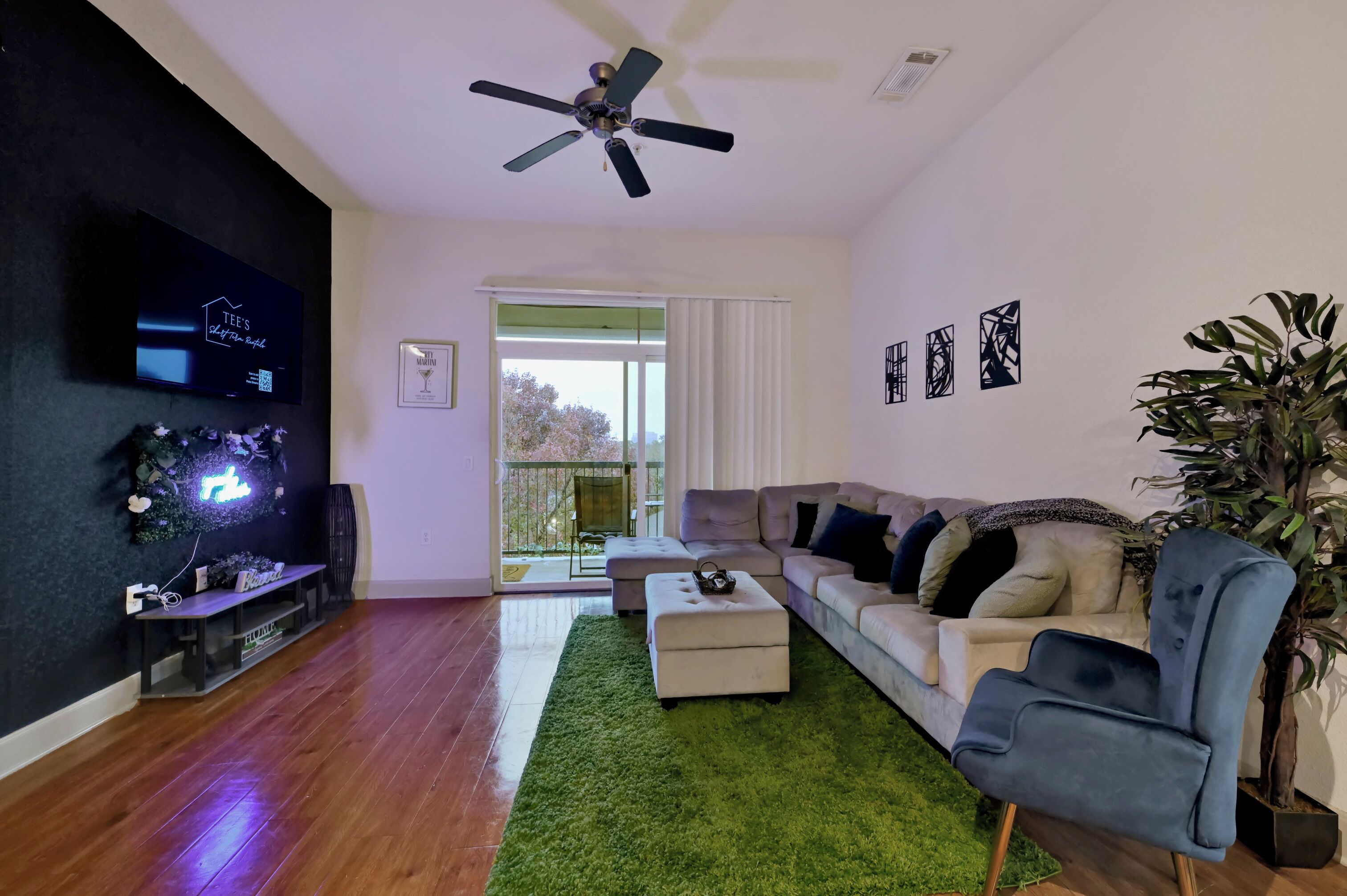 A living room with a flat-screen TV, ceiling fan, sofa, ottoman, and a large plant.