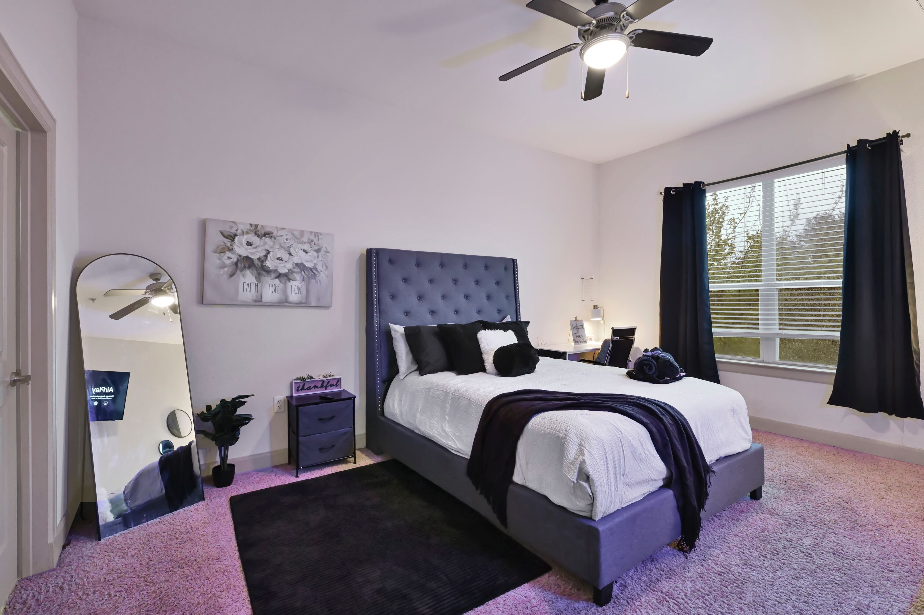 Een slaapkamer met een bed, een plafondventilator, een raam met gordijnen, een spiegel en een klein tafeltje met een plant.