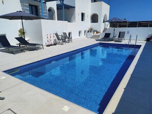 Une piscine rectangulaire à l’eau d’un bleu cristallin, entourée d’un bâtiment blanc agrémenté de touches de bleu et dotée d’un espace de détente extérieur.