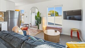 A modern living room with a sofa, a round coffee table, a TV mounted on the wall, and a kitchen area with a refrigerator. - 10 Mins To Joshua Tree National Park & Marine Base. Fire Pit, Romantic Retreat (Twentynine Palms)