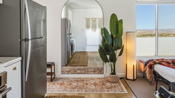 A modern kitchen with a stainless steel refrigerator, a dining area with a table and chairs, a large potted plant, and a view of the desert through a large window. - 10 Mins To Joshua Tree National Park & Marine Base. Fire Pit, Romantic Retreat (Twentynine Palms)