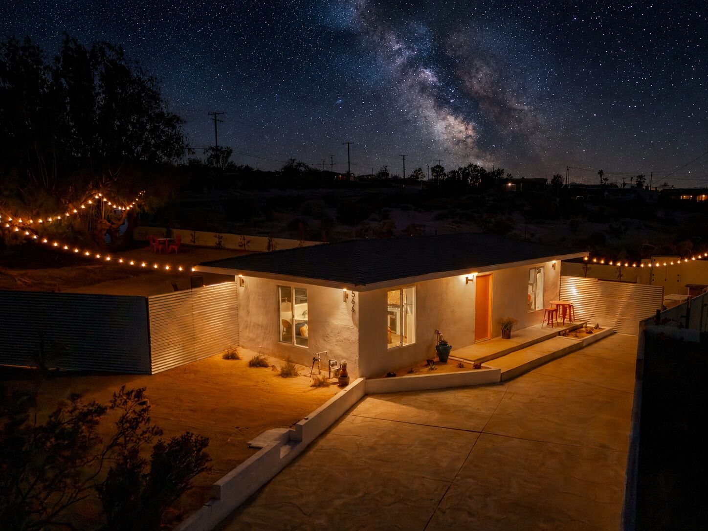 Une maison moderne offrant une vue dégagée sur la Voie lactée, un patio illuminé de guirlandes lumineuses et une entrée bien éclairée.