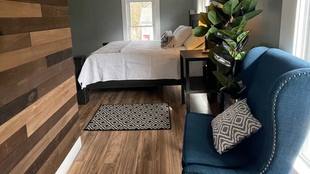 A bedroom with a bed, a blue armchair, a ceiling fan, a potted plant, and a window. - Green Mountain Retreat (Rutland)