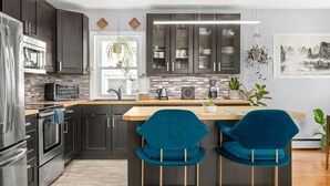 A modern kitchen with dark cabinets, a stainless steel refrigerator, a built-in oven, and a microwave. There are two blue bar stools and a wooden island with a sink. - ✪ Ramsay Manor - Walk to Stampede in 10 mins ✪ (Calgary)