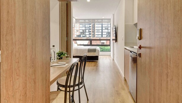 Un interior moderno con zona de comedor, cama y vistas a edificios a través de amplios ventanales.