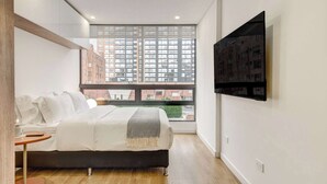 Un dormitorio moderno con un ventanal grande, una cama, una mesa redonda y un televisor montado.