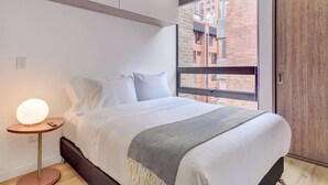 Una camera da letto moderna con un'ampia finestra, un letto con lenzuola bianche, un comodino con una lampada e vista su un edificio in mattoni.