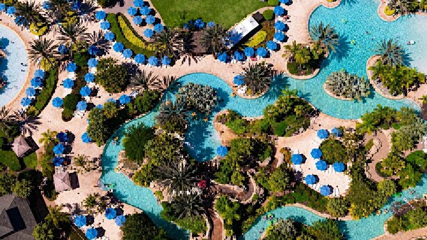 Eine Vogelperspektive zeigt ein Resort mit mehreren Swimmingpools, Liegebereichen mit Sonnenschirmen und üppiger Bepflanzung.